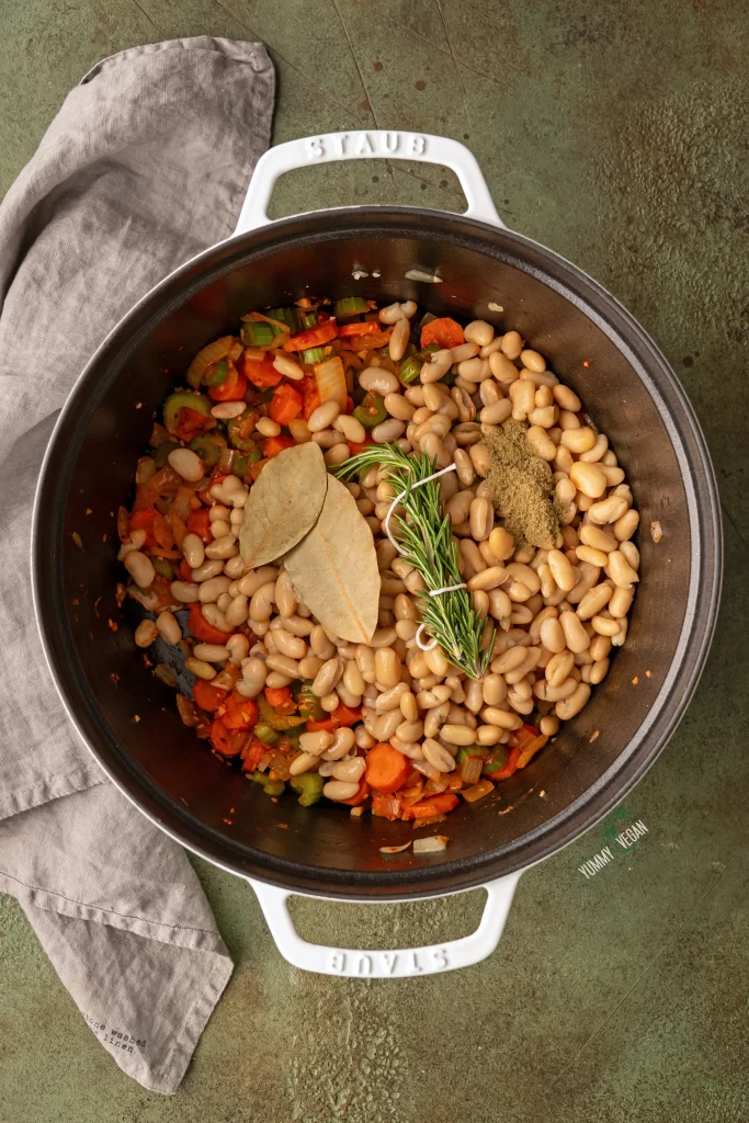 bay leaves and fresh herbs added to a soup pot with beans and veggies